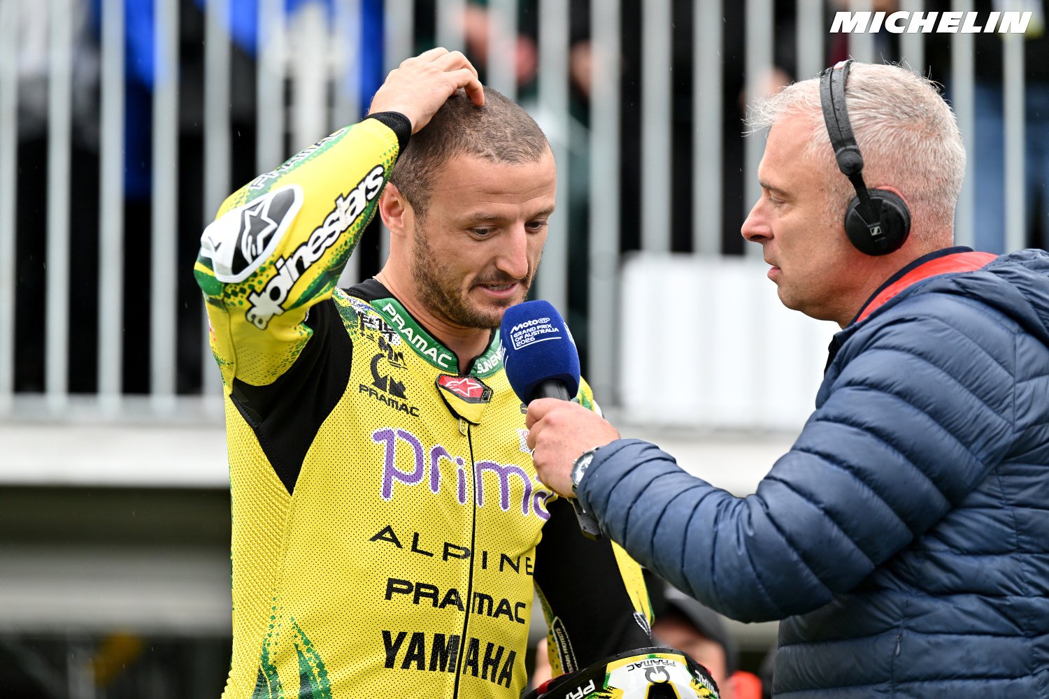 A rider in yellow Pramac Yamaha MotoGP leathers touches his hair while a reporter with headphones holds a microphone to him in the paddock