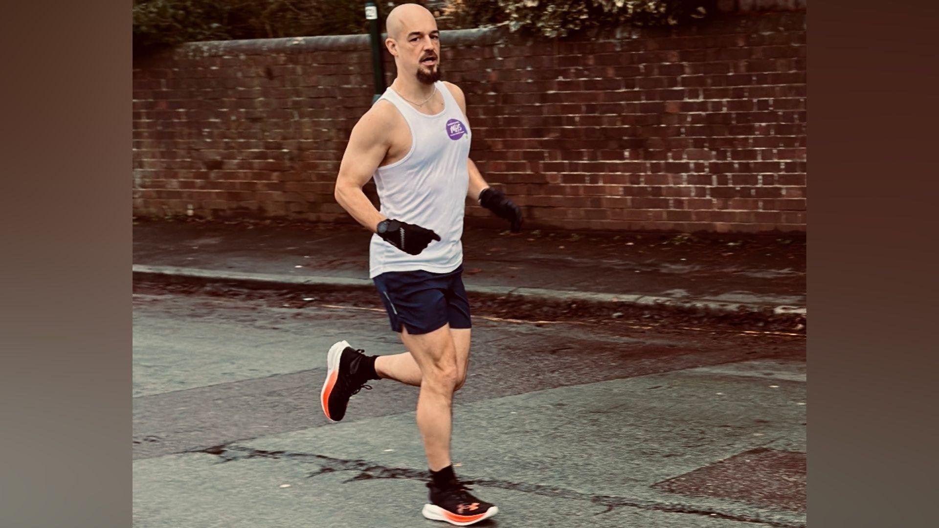 A bald man with a white vest and black shorts is running along a rainy road.