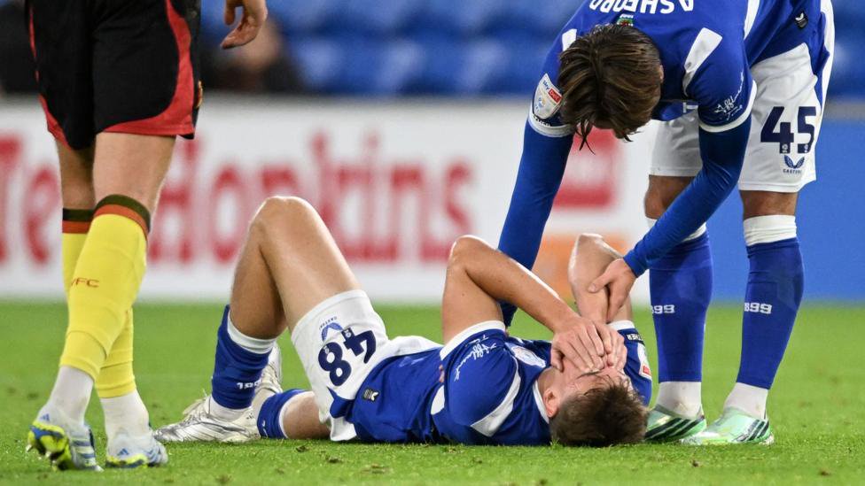A team-mate consoles Dylan Lawlor who lies on the pitch in his blue and white Cardiff City kit covering his face with his hands.