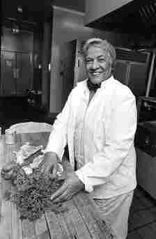 A black and white photo of a middle-aged woman in a chef's coat in a professional kitchen with herbs and vegetables
