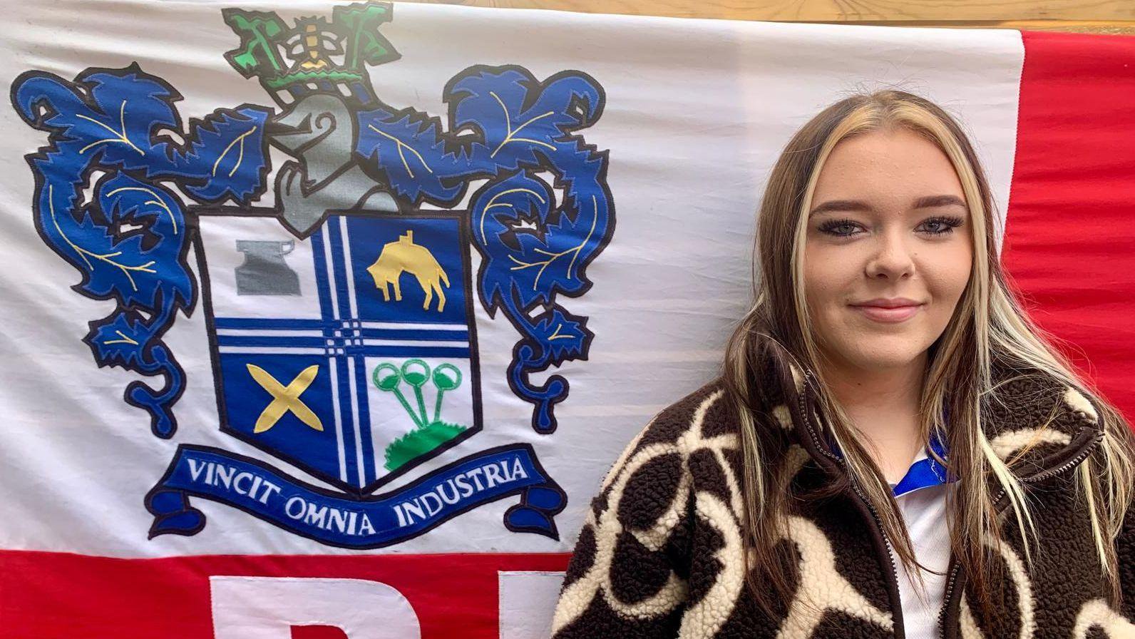 Katie Maken stands in front of a large Bury FC flag displaying the club crest and the motto “Vincit Omnia Industria”. She has long brown and blonde hair.