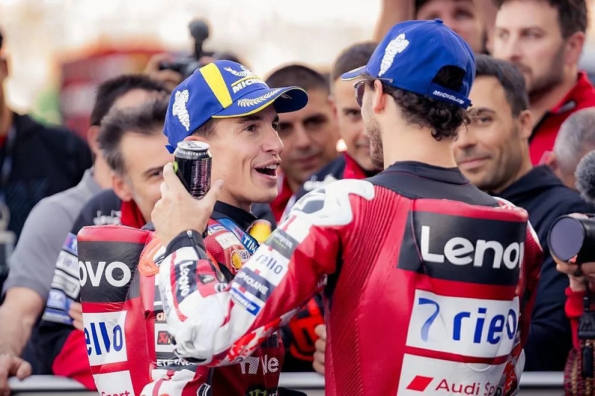 Two riders in red Ducati leathers and blue caps chat in a crowded paddock; one holds a drink can, with Lenovo and Audi sponsor logos visible.