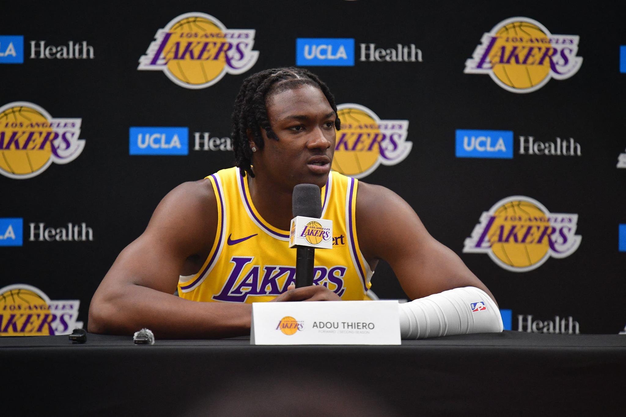 Los Angeles Lakers player Adou Thiero speaks into a microphone during media day, sitting behind a table with a nameplate in front of a backdrop featuring Lakers and UCLA Health logos.