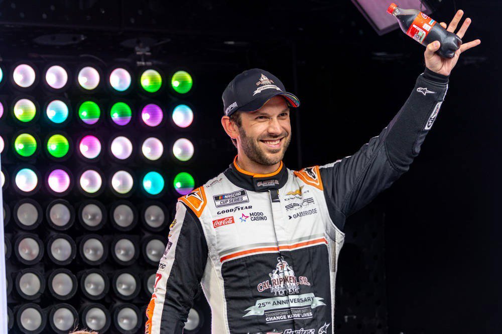 Race driver in a black and white firesuit with orange trim and a Cal Ripken Jr. Foundation 25th anniversary patch, smiling and raising a hand by a stage with…
