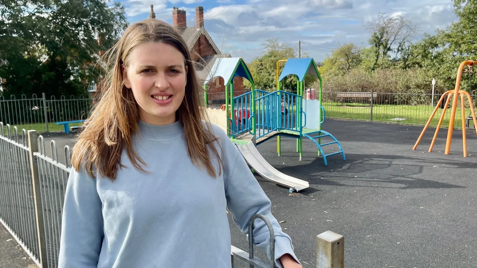 BBC A woman with long hair is stood by a children's playground complete with a brightly coloured climbing frame and swing set.