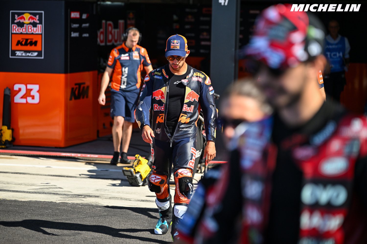 Rider in Red Bull KTM leathers walks through the MotoGP pit lane past the KTM garage, with a blurred rider in foreground