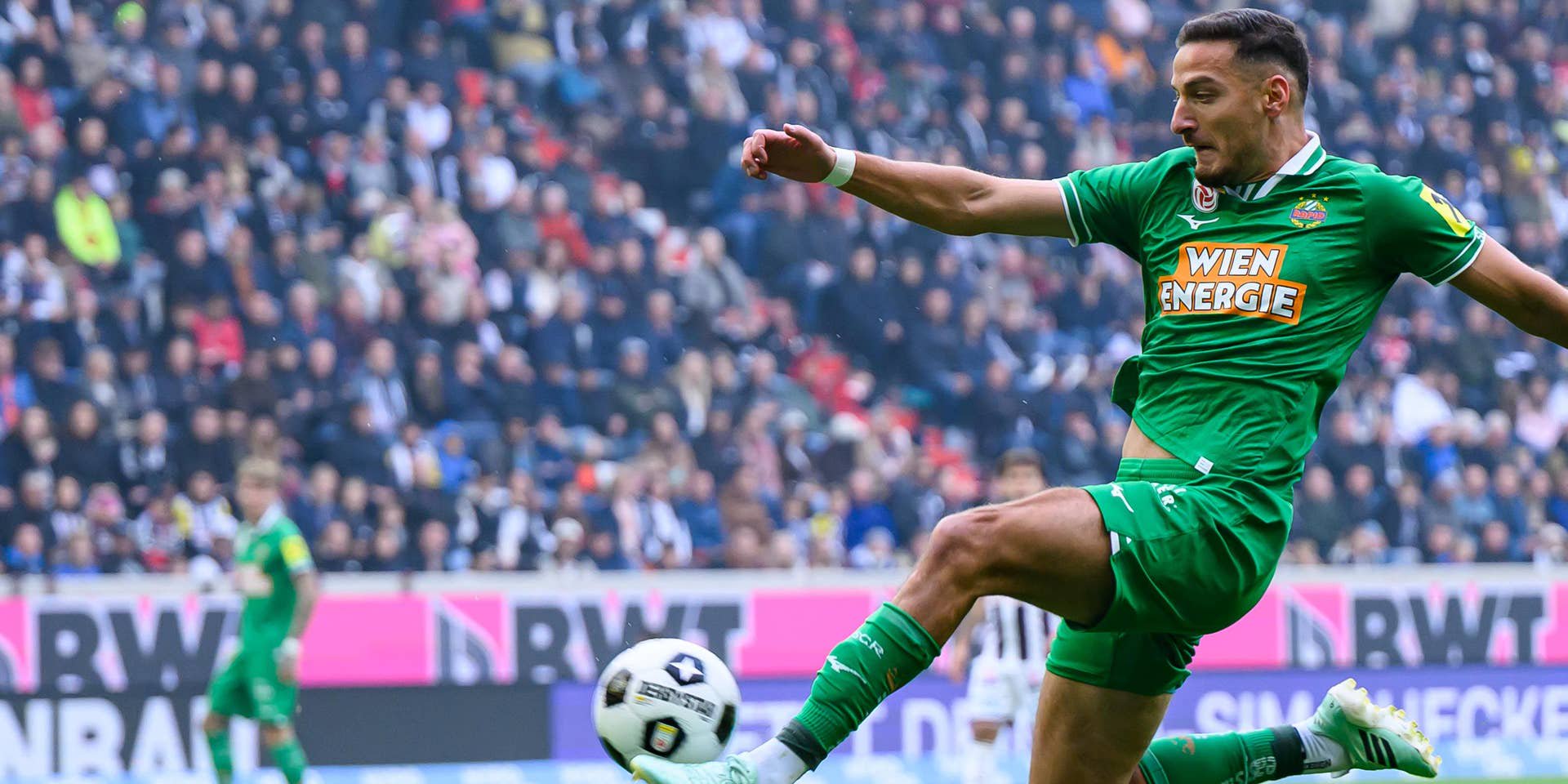 Ein Fußballspieler von Rapid Wien springt und schießt den Ball während eines Spiels im Stadion.