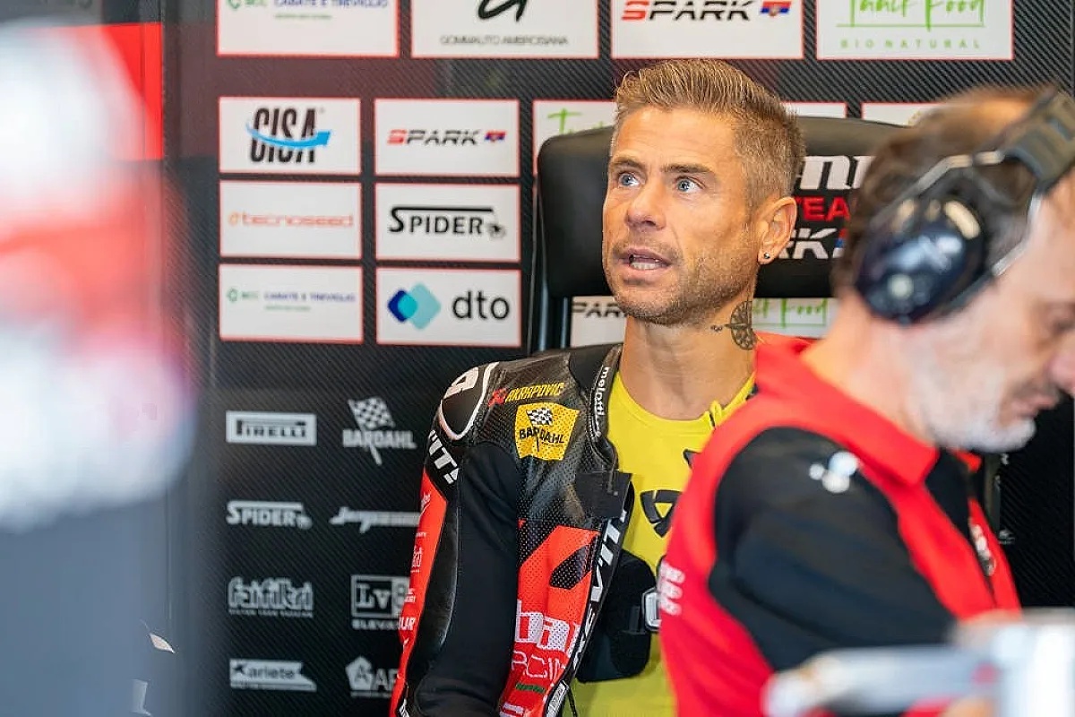 A rider in a black and yellow shirt and Ducati-style leathers in a garage, with sponsor banners behind him and a team member in red in the foreground