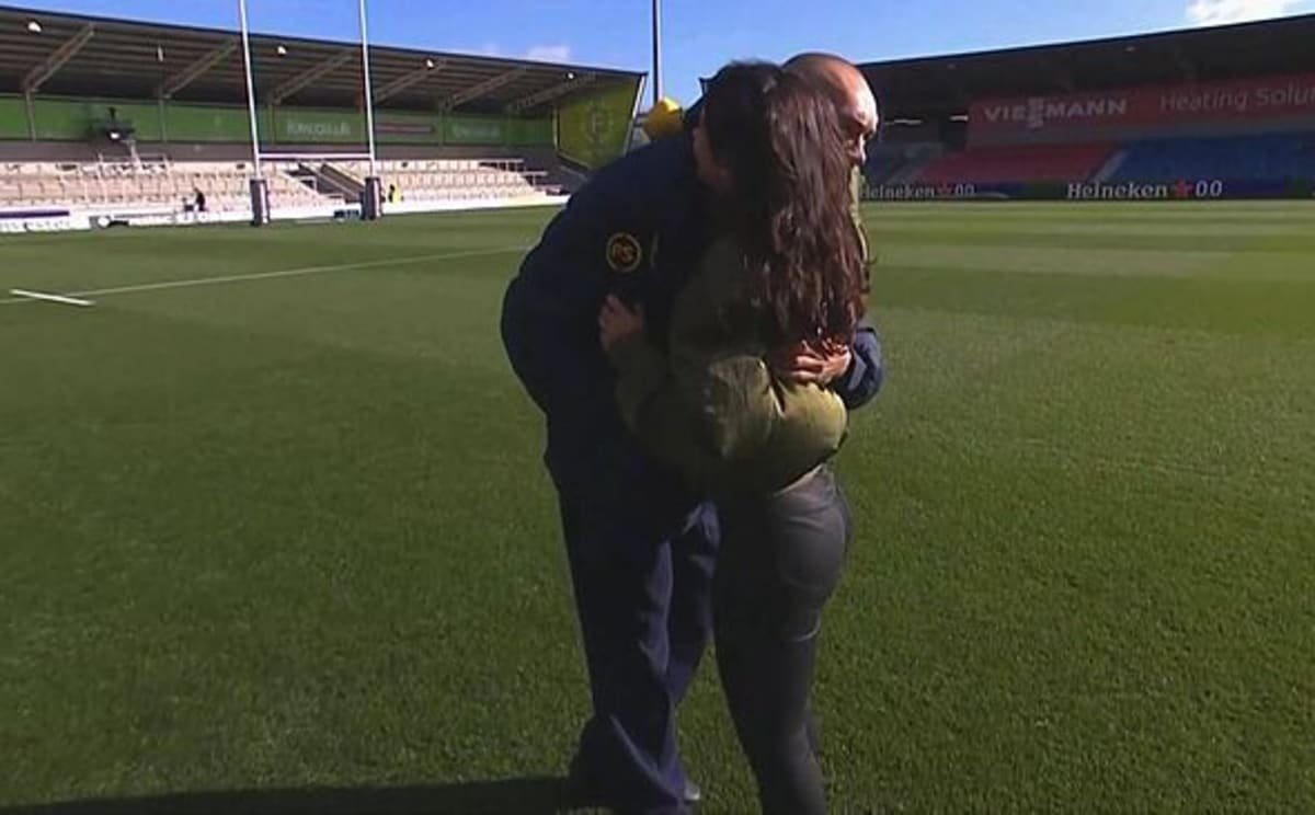 An individual is standing on a wide open sports field, holding a ball, with their back turned to the camera, and an empty stadiu