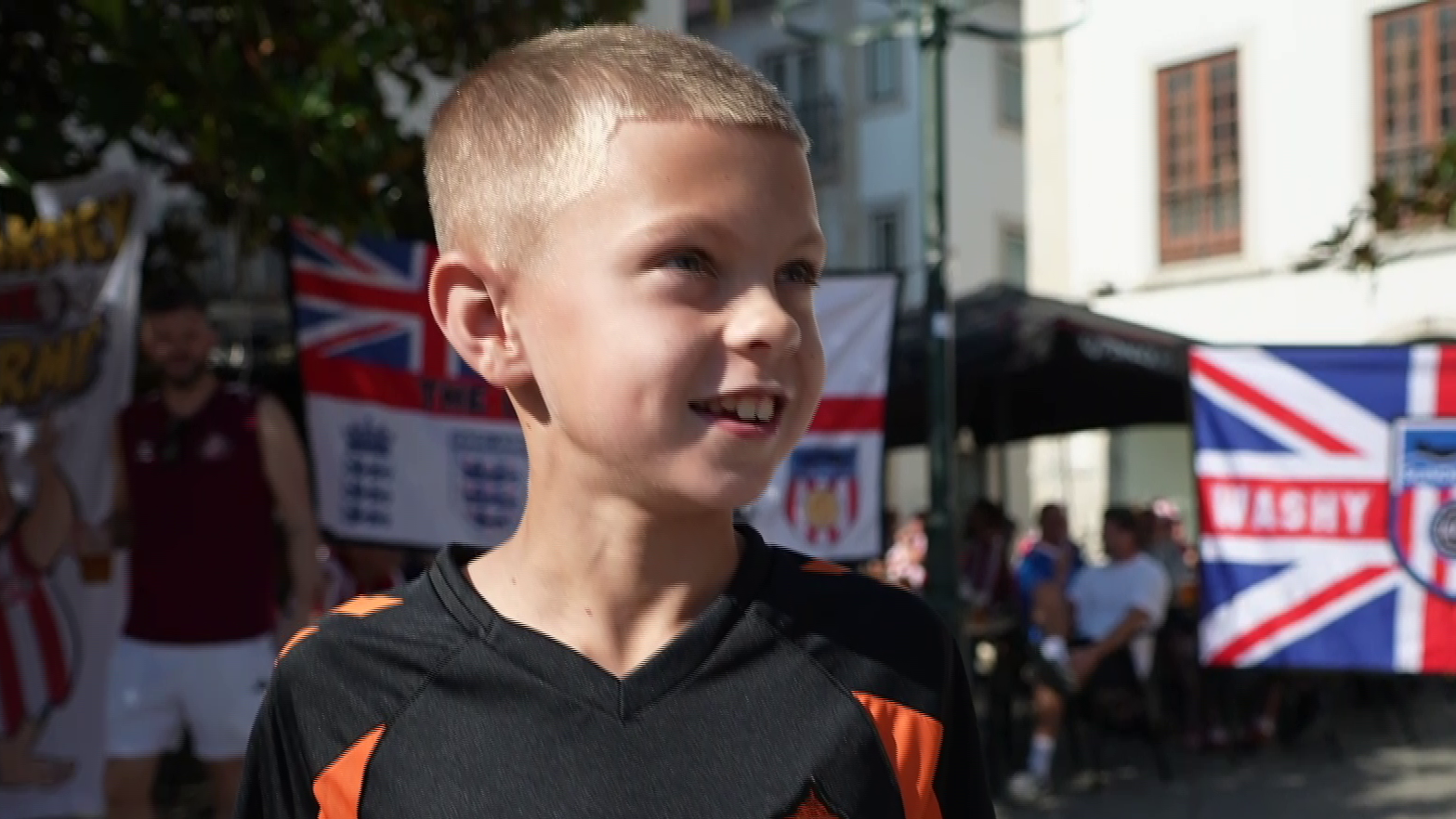 George has short-cropped blond hair and is smiling at the camera. He is wearing a black football top with orange highlights. He is standing in a sunny square and there are Union Jacks in the background as well as other football fans. 