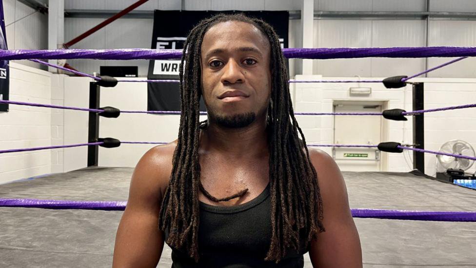 A 28-year-old man with long dreadlocks looks into the camera. He has a stern expression and is wearing a black vest. Behind him is a wrestling ring.