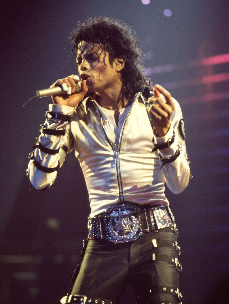 Michael Jackson performs on stage in a white jacket and black trousers, holding a microphone in his right hand.
