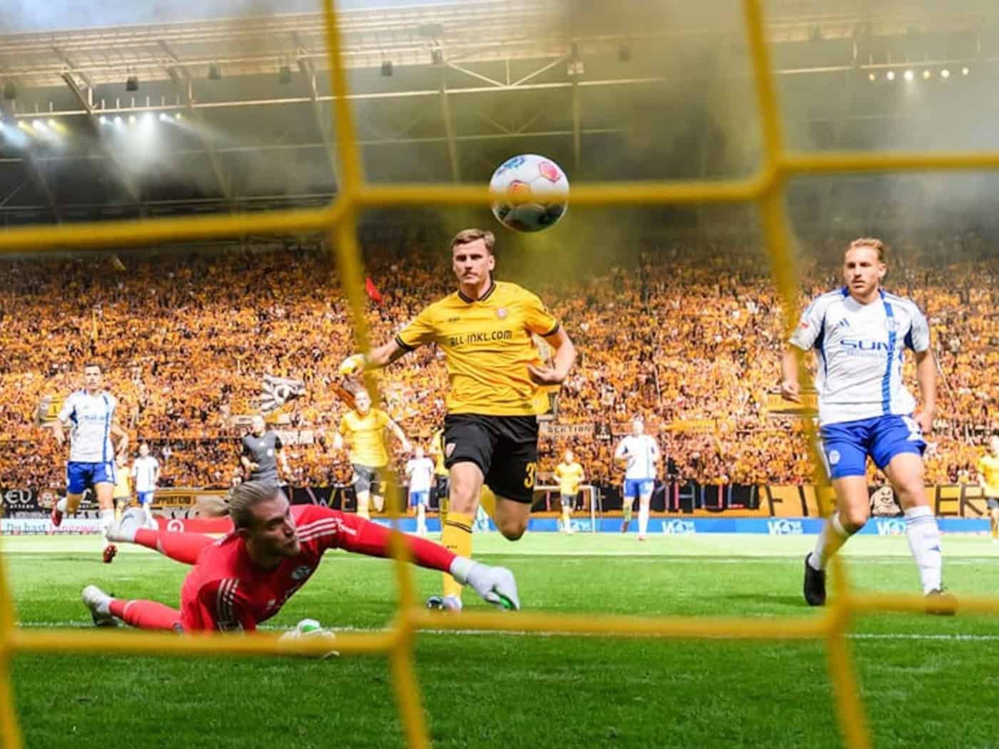Blick durch das Tornetz auf eine Spielszene im voll besetzten Rudolf-Harbig-Stadion: Ein Dynamo-Spieler in Gelb köpft den Ball im Strafraum, während der Schalke-Torwart am Boden liegt. Auf den Rängen der gefüllten Fan-Tribüne steigt im Hintergrund Rauch auf.
