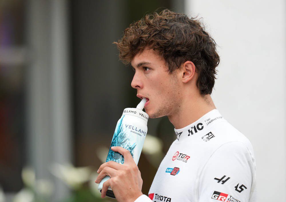 A driver in a white Toyota Gazoo Racing suit drinking from a Vellamo water bottle in the F1 paddock, with Toyota and TGR logos visible