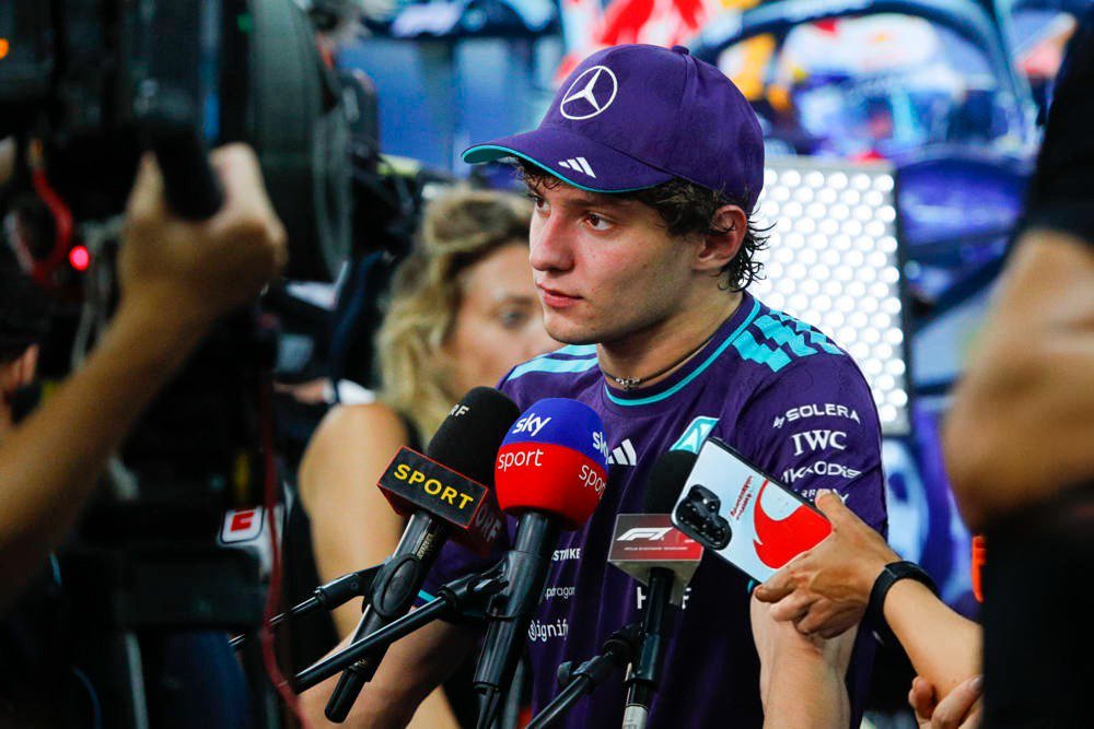Driver in a purple Mercedes cap and team shirt speaking to reporters, with Sky Sport and F1 microphones in front of him at a paddock
