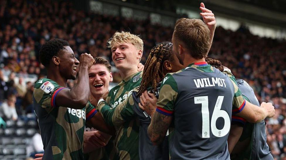 Stoke City defender Luke Graham, in camouflage kit, smiles broadly as he is surrounded by team-mates, all celebrating his goal at Derby County