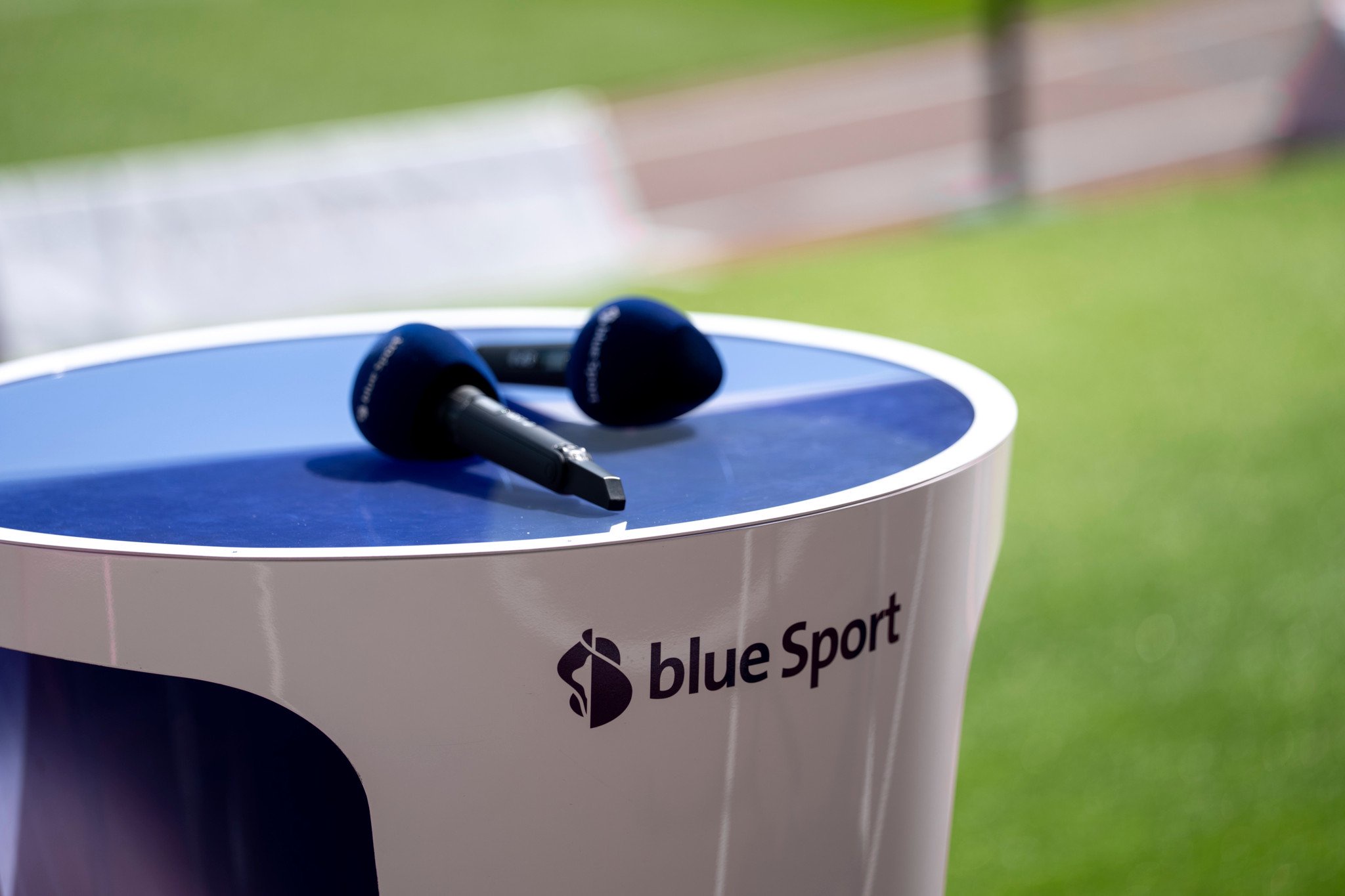 Zwei Mikrofone liegen auf einem weiss-blauen Moderationspult mit dem Logo von Blue Sport, im Hintergrund grüner Rasen.