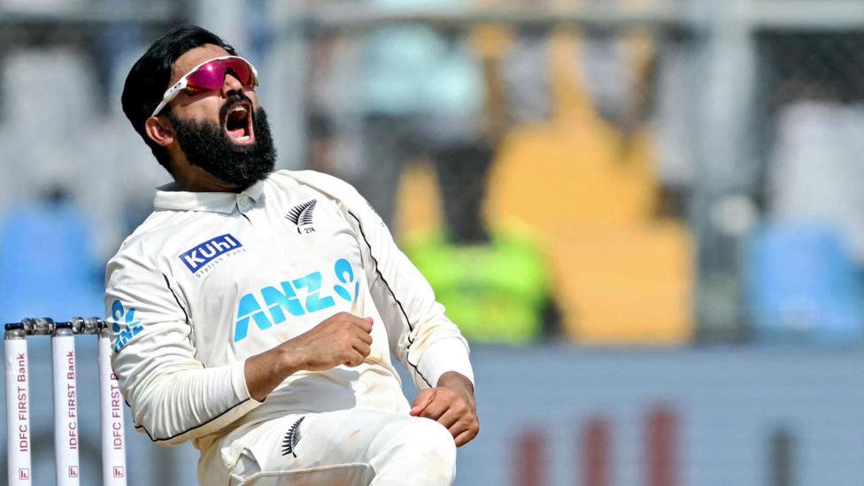 New Zealand spinner Ajaz Patel with sunglasses on and down on one knee as he celebrates victory against India in front of the wickets