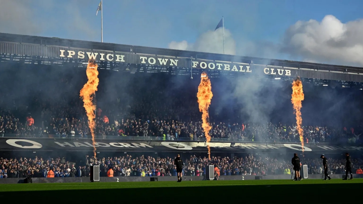 Tony O'Brien/Reuters A two-tiered stand at a football ground. It has "IPSWICH TOWN FOOTBALL CLUB" in white letters on the front of the roof. Between the upper and lower tiers is a sloping roof over executive boxes which has advertising on it. There are three flames rising from devices on the pitch that reach the roof of the stand. The stand is full of supporters.