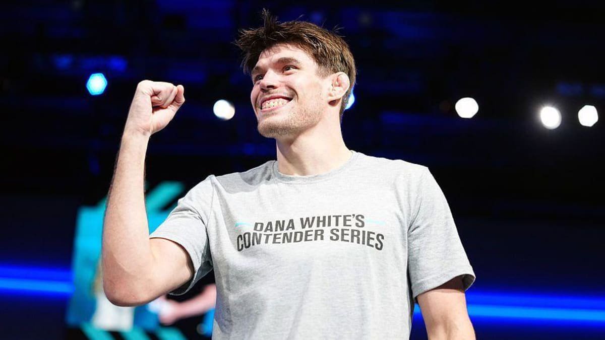 A man stands on a brightly lit stage wearing a grey T-shirt with text saying “DANA WHITE’S CONTENDER SERIES.” He is raising his fist and smiling towards a crowd of people off-camera. Blue stage lighting and several bright spotlights are visible in the blurred background.