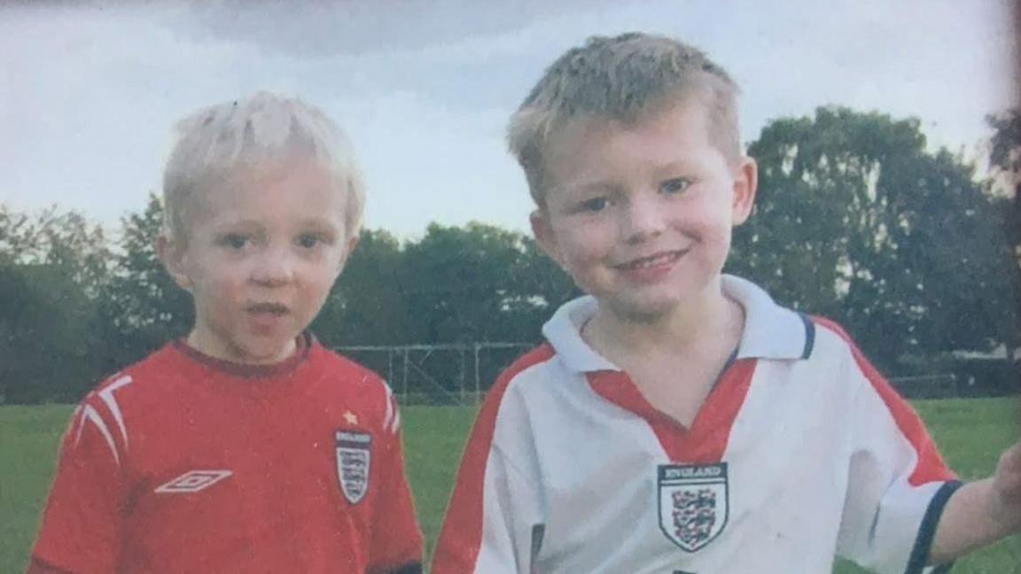 An old photo of two young boys on a football pitch. The boy on the left is wearing a red England football shirt and the one on the right is wearing a white England football shirt. 