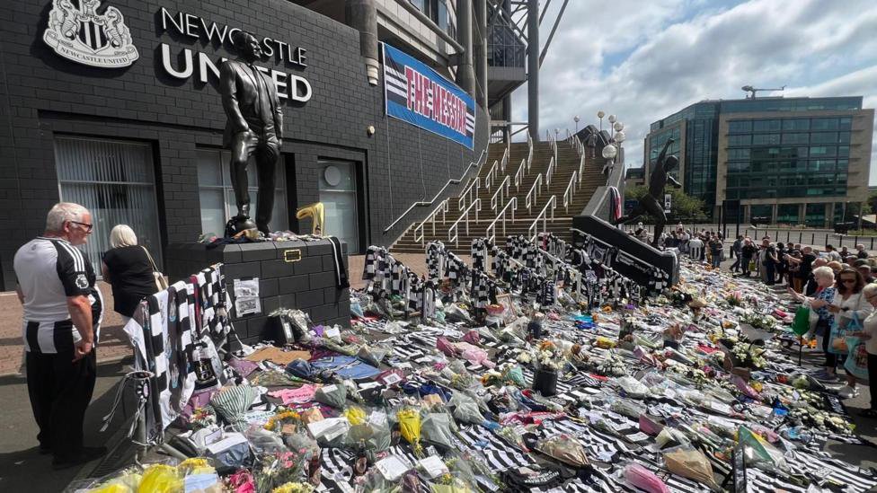 People lined up looking at the tributes to Kevin Keegan on the floor outside St James' Park in Newcastle, including flowers, cards and football shirts. There is a blue and white banner hanging from the side of the stadium, between the Newcastle United sign and the steps, with the words The Messiah - in recognition of Keegan.