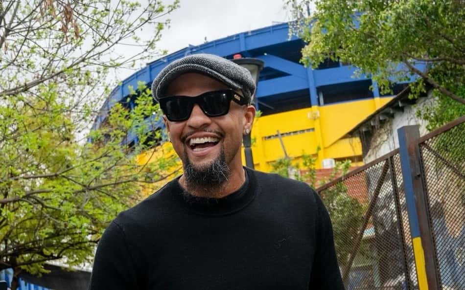 Felipe Melo com boné cinza, óculos escuros e casaco preto sorri ao ar livre, com grade e estrutura azul/amarela ao fundo