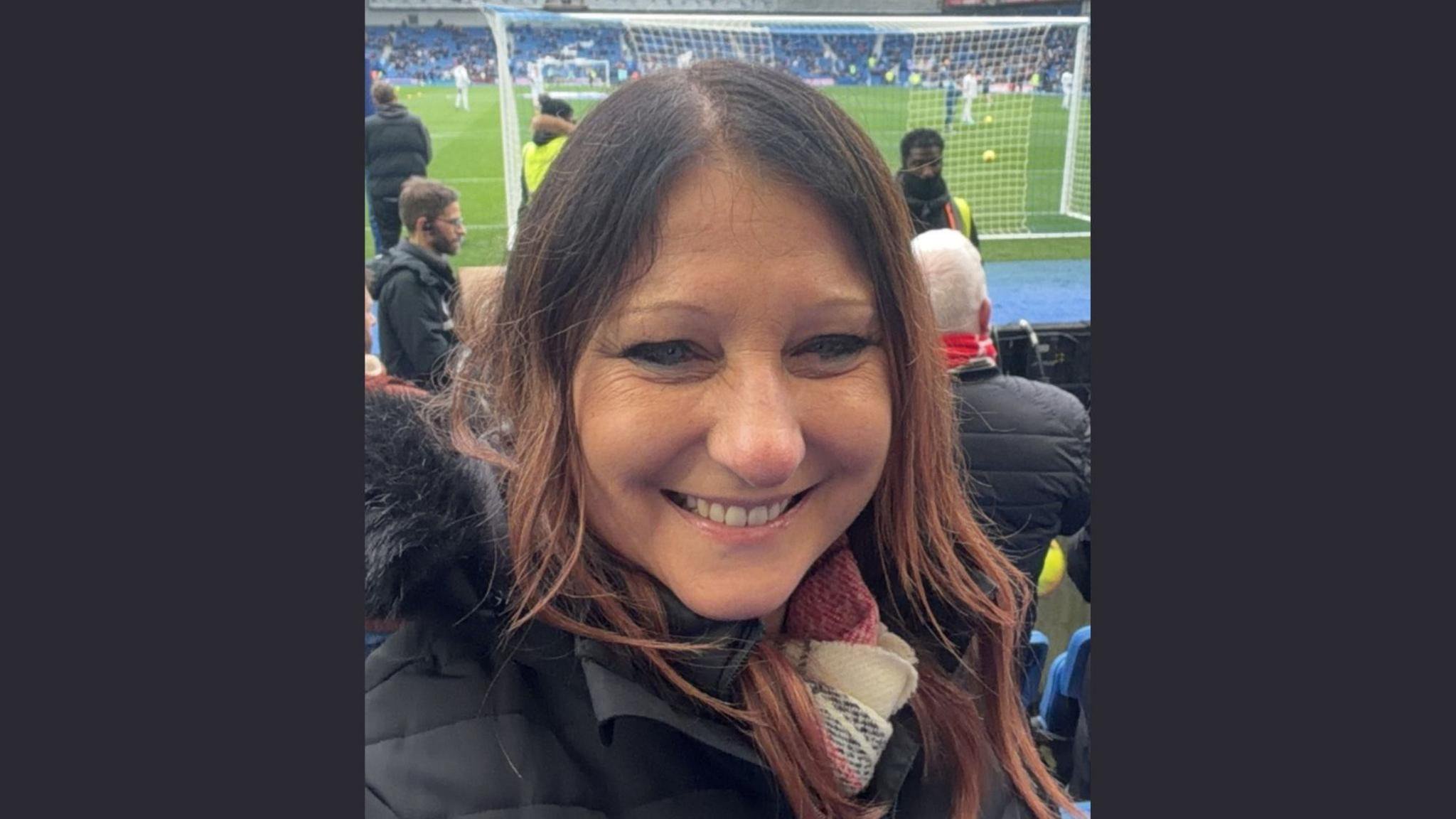 Picture of a white female with brown hair, wearing a scarf and black coat, pictured in front of a football pitch.