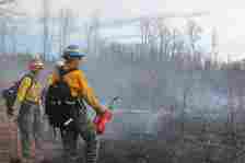 Two women firefighters stand in a burned field, carrying a blow torch and a hand tool.