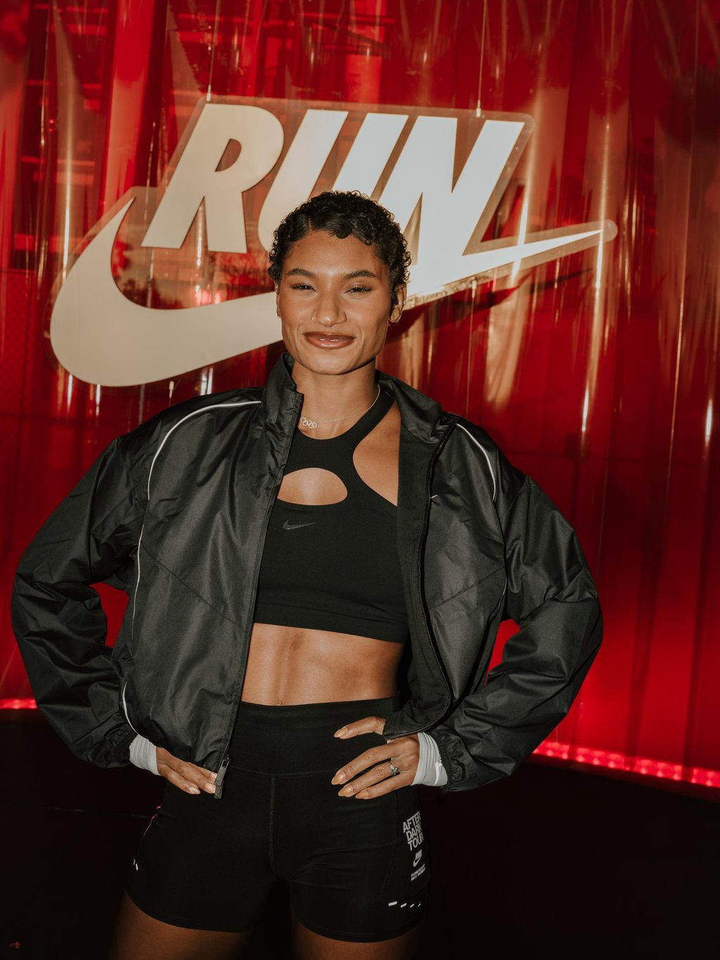 A woman poses in front of a Nike sign, wearing a black sports bra and jacket.