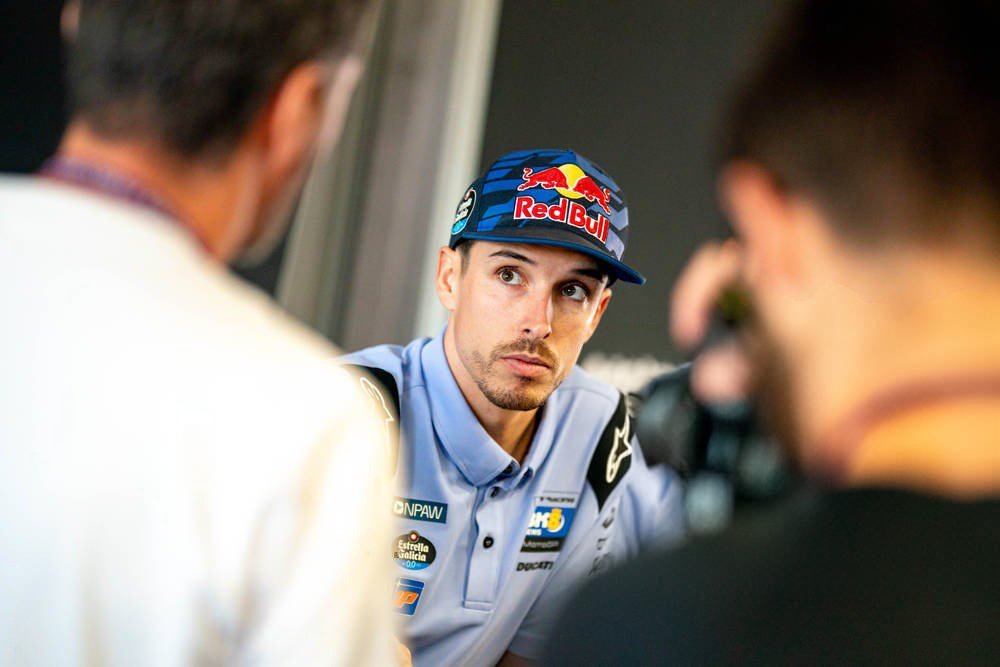 A MotoGP rider in a blue Red Bull cap and light blue team polo with Ducati and sponsor logos, speaking intently during a press interview in the paddock