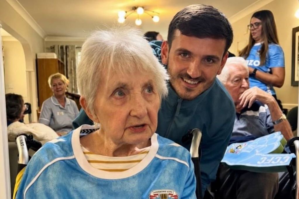 An elderly woman wearing a blue jersey is standing next to a smiling man, both of them posing for a photograph in a room with several other individuals in the background.