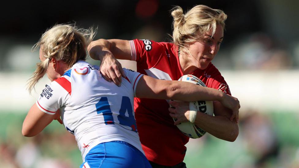 Bethan Dainton in the red of Wales runs with the ball as she tries to evade a tackle from a white-shirted England player.