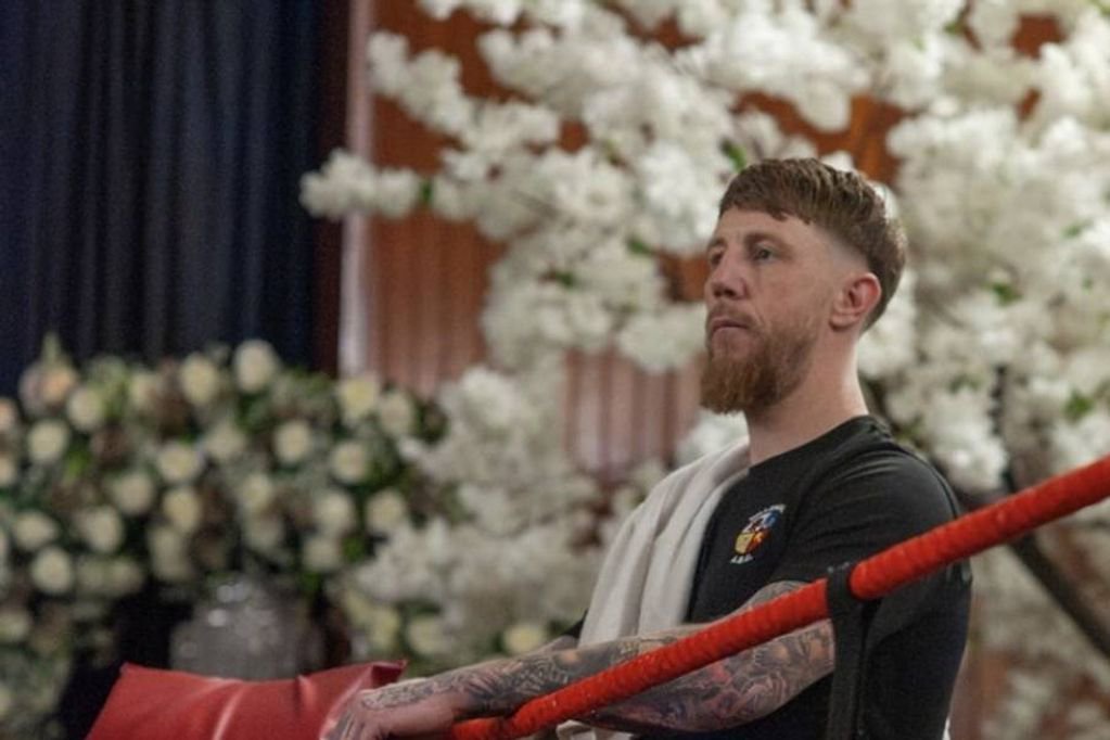 An individual with a beard, sporting tattoos on his arms, is holding a red boxing glove, standing against a backdrop adorned with white flowers.