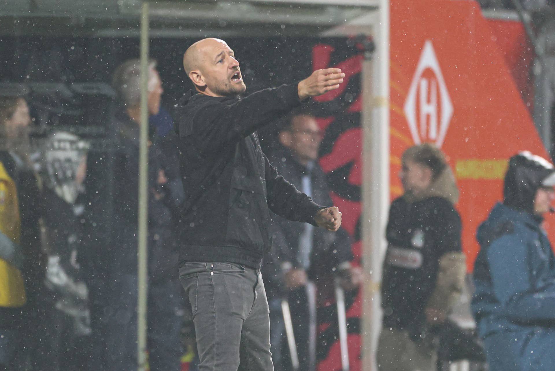 Ein Trainer gestikuliert am Spielfeldrand während eines Fußballspiels im Regen.