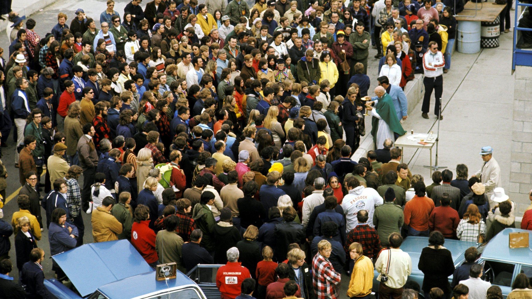 A priest conducts a church service in the pits at the United States Grand Prix at Watkins Glen in 1973. A large crowd is gathered in front of him.