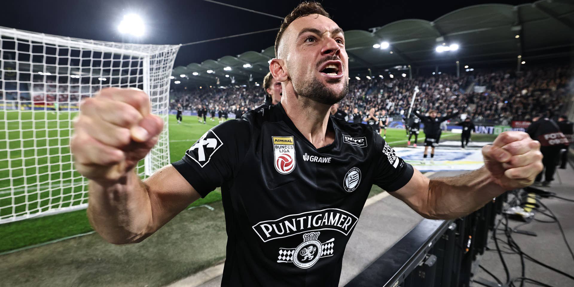 Ein Sturm-Graz-Spieler jubelt im schwarzen Trikot mit geballten Fäusten und lautem Schrei nah vor der Tribüne im Stadion.