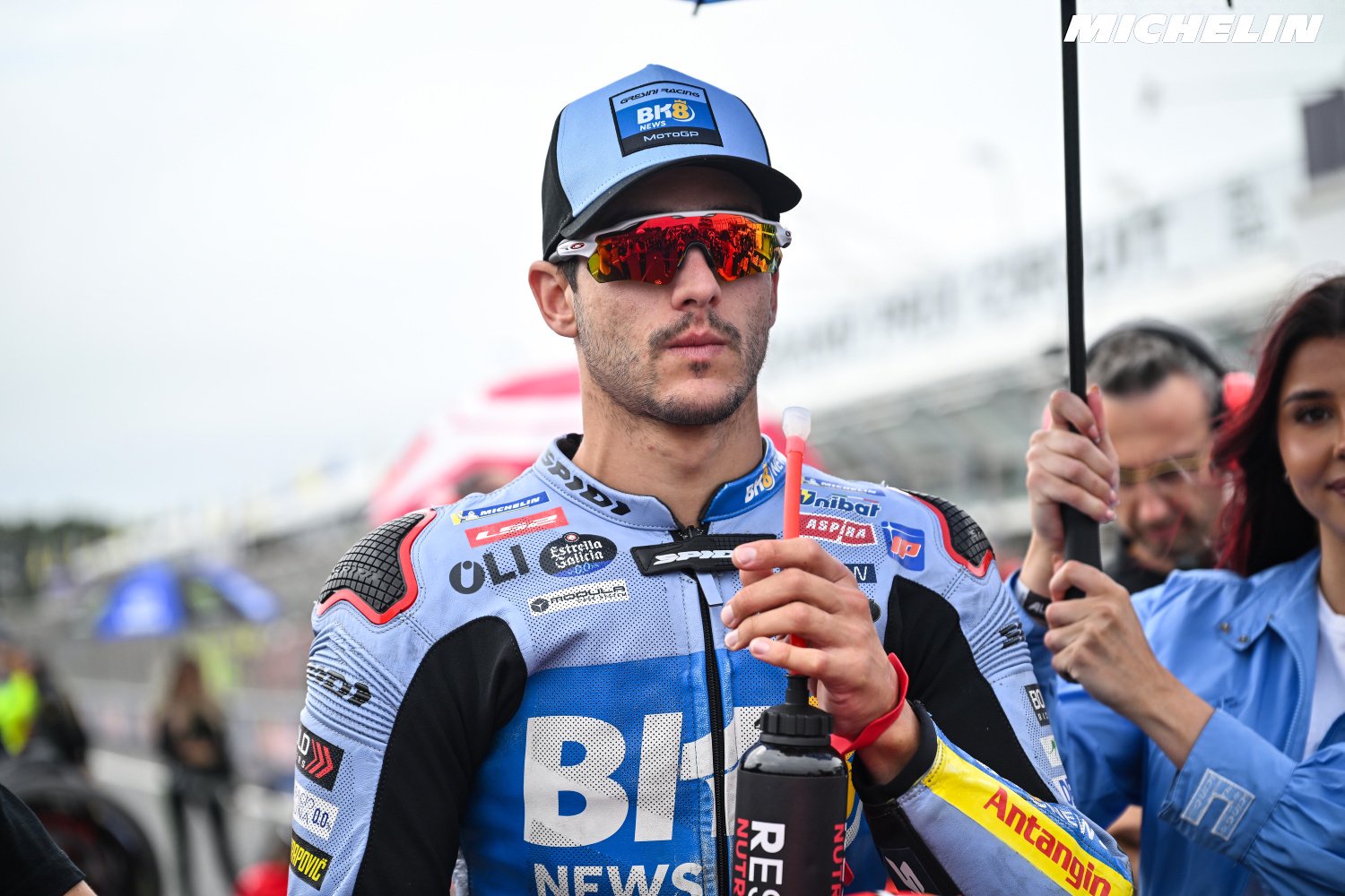 Rider in blue and light-blue leathers with BH Motor cap and red-tinted sunglasses on the MotoGP grid, Gresini-style livery, Michelin branding behind