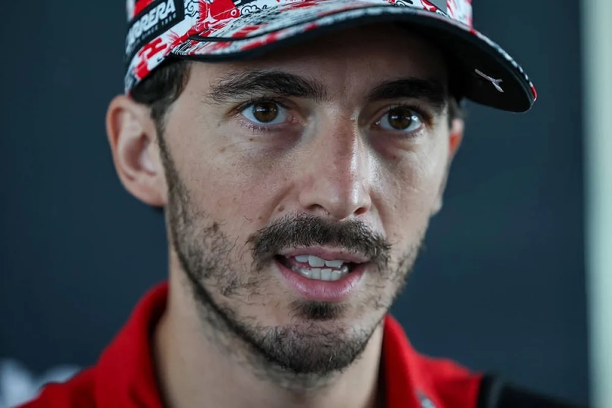 A Ducati rider in a red cap with a red-and-white patterned design and red collared shirt speaks to reporters in the paddock.
