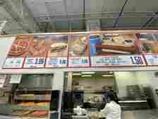 Restaurant menu board showing various food items with prices, including chicken bake, roast beef sandwich, hot dog combo, and pizza. Staff member in foreground
