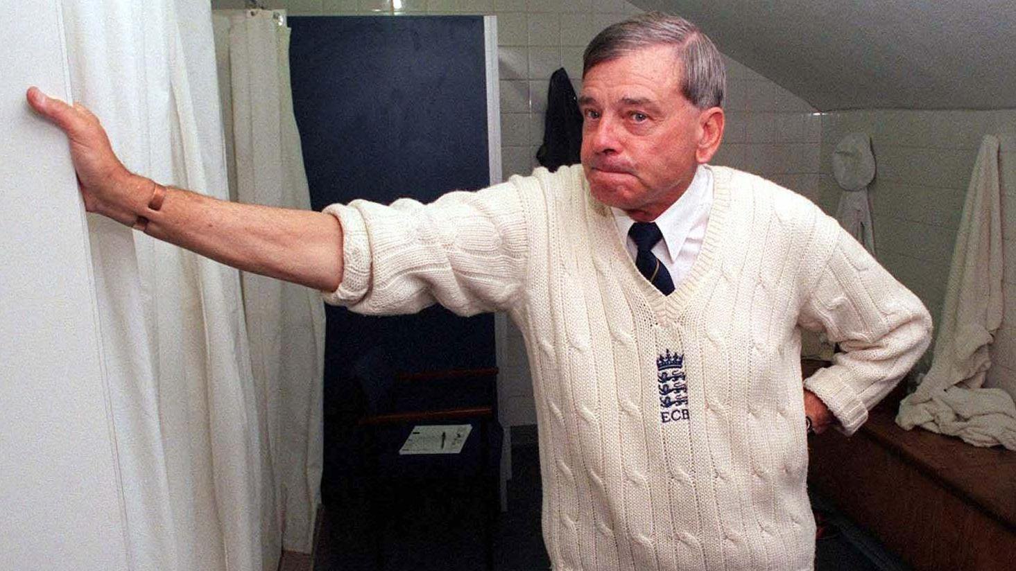 Bird stands in a back room, with his arm rested on the wall. He is wearing a traditional cricket vest, branding with the ECB logo stitched. He wears a white shirt and tie underneath.