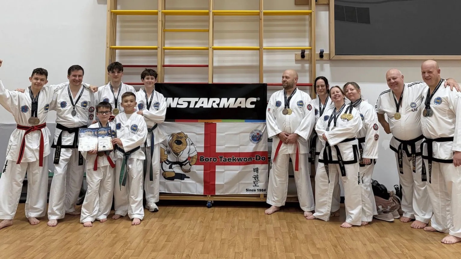 Isabella Verona/BBC A group of taekwondo club members pose for a photograph inside a sports hall. Several participants wear medals around their necks and stand behind a club banner and flag. The group includes both adults and children dressed in white ITF taekwondo uniforms.