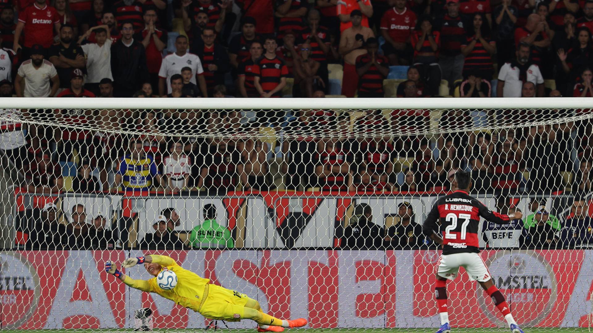 Goleiro de uniforme amarelo se estica para defender uma bola, enquanto um jogador de camisa preta e vermelha com o número 27 observa. Torcedores lotam as arquibancadas ao fundo.