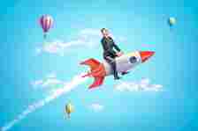 Young businessman riding toy rocket in blue sky with hot air balloons in background.