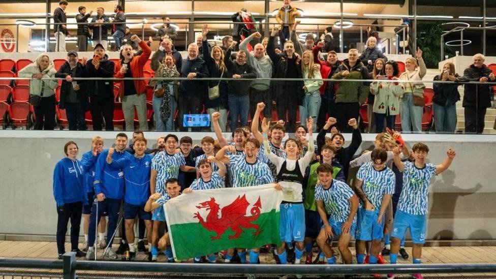 Cardiff Met U19s in blue and white shirts celebrating holding a Welsh flag with their friends and family above them in a stand