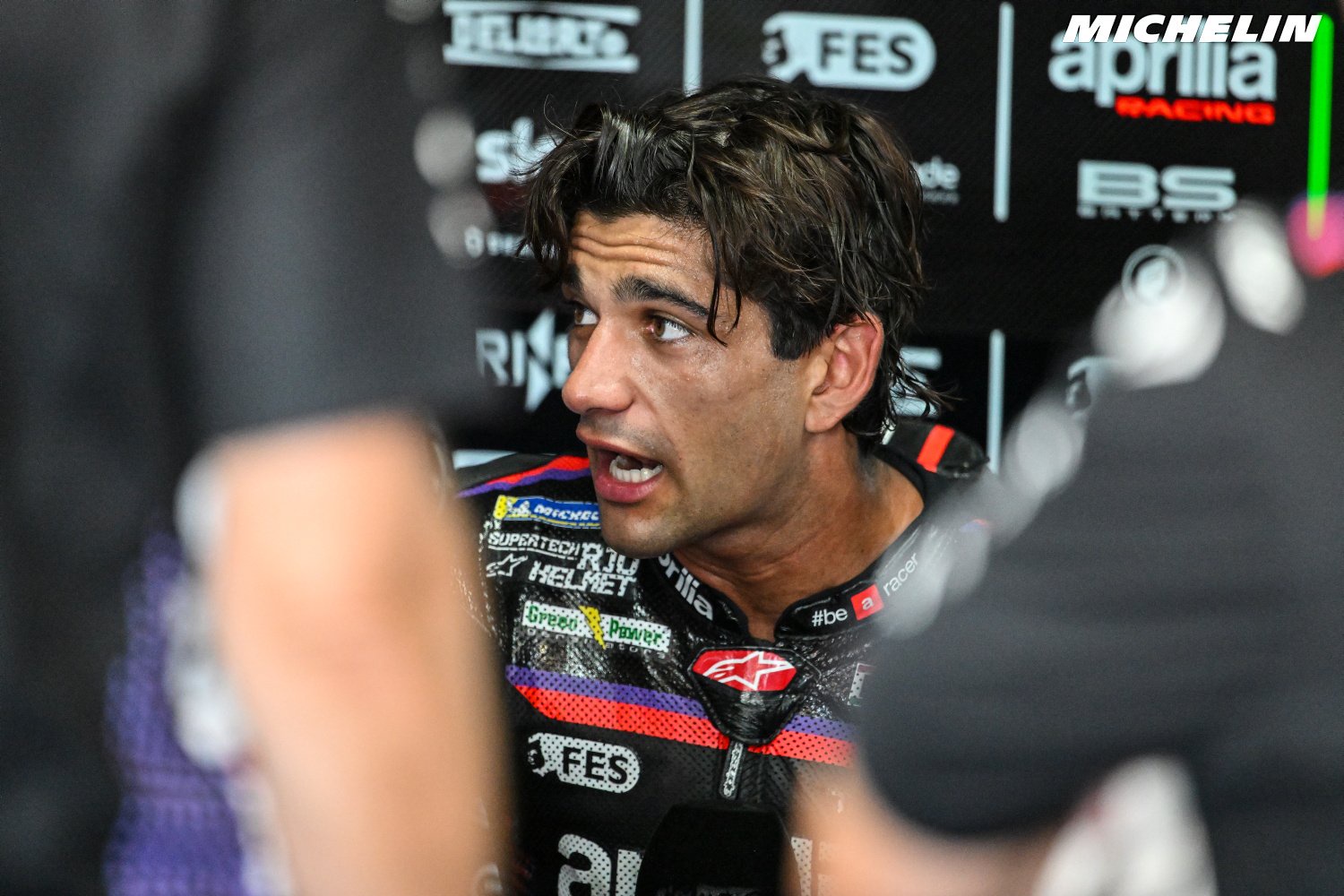 Rider in black Aprilia Racing leathers speaks intently in the MotoGP paddock, with team sponsor logos behind him and blurred figures in the foreground.
