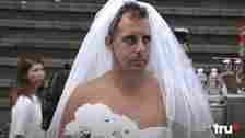 Joe Gatto, wearing a wedding veil and strapless top, holding a bouquet of white flowers. Stairs and bottles in the background. TruTV logo at bottom right
