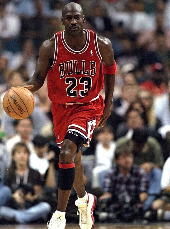 Basketball player in a red Bulls jersey, number 23, dribbles the ball on a court. The player wears a red wristband and white sneakers, and is in motion during a game, with a blurred crowd in the background.