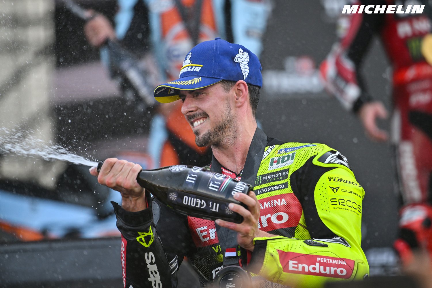 Rider in a blue Michelin cap and lime-yellow Enduro leathers sprays champagne from a bottle on the MotoGP podium