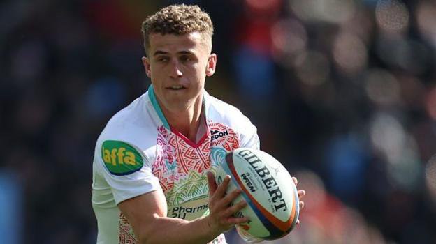 Billy Searle in action for Leicester Tigers. He is holding a rugby ball and is poised to pass it to his right. He is wearing a white shirt with red and green patterning. 